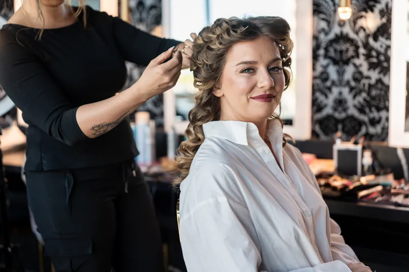 A stylist pins up Lauren's braided updo while she sits in a white robe during wedding day preparations.