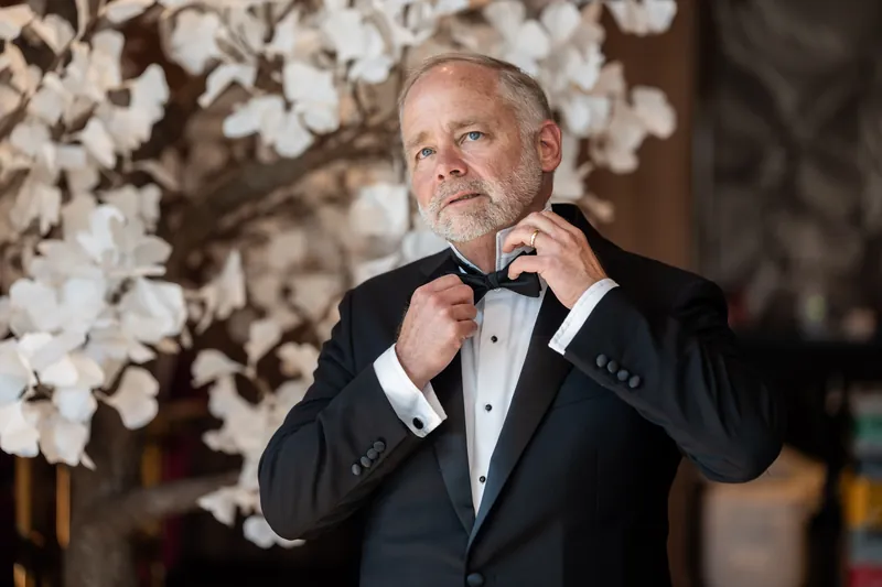 Kevin adjusts his black tuxedo bow tie in front of a backdrop of white flowering branches.