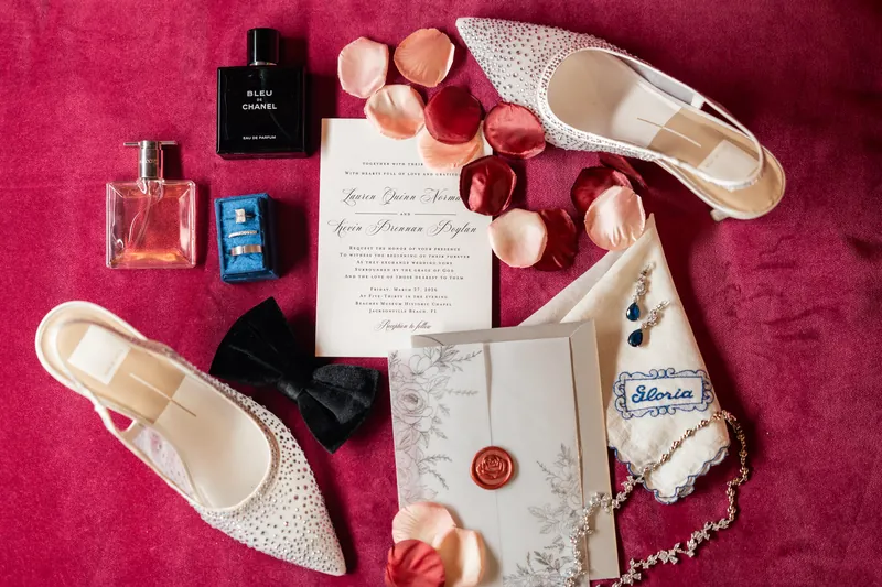 Wedding day details arranged on magenta fabric include ivory heels, perfume bottles, invitations, rose petals, and jewelry.