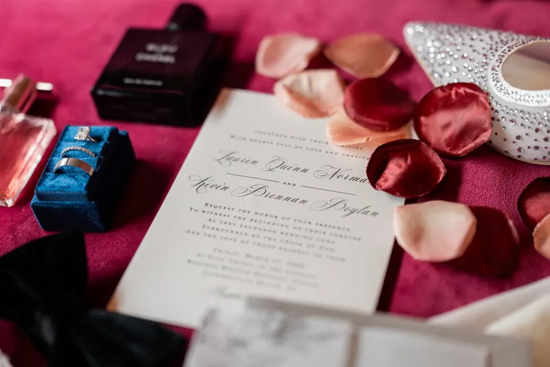 Close-up of Lauren and Kevin's wedding invitation surrounded by cosmetics, candles, and rose petals on a pink background.