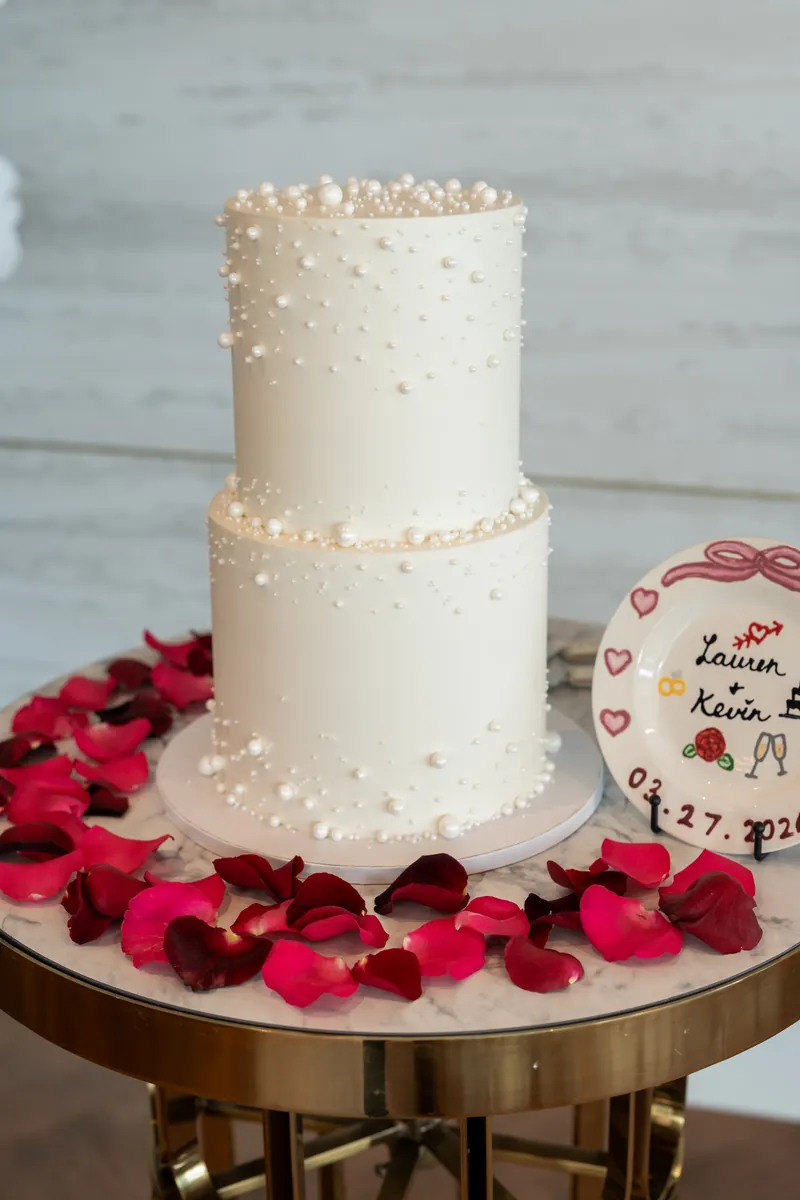 A two-tier ivory wedding cake decorated with pearl beads sits on a gold stand surrounded by red rose petals.