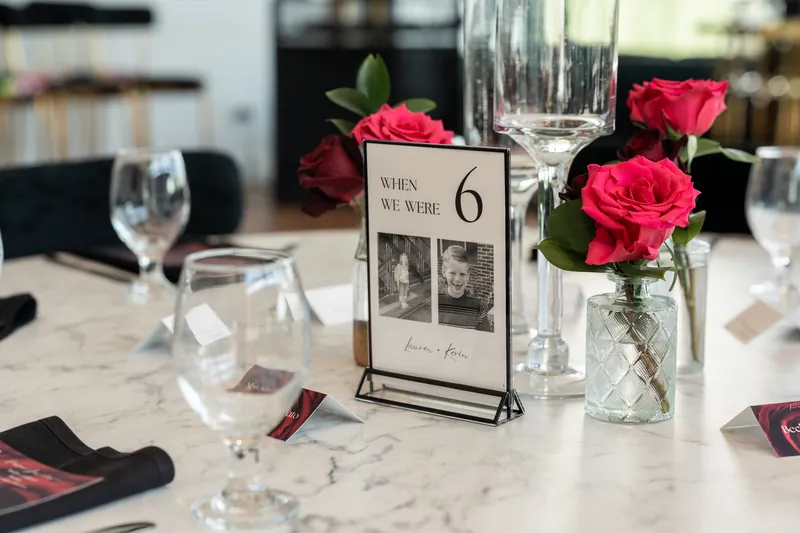 Marble table with table number card, pink and red roses in glass vases, wine glasses, and place settings