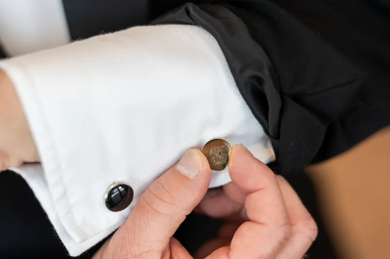 A groom in a black suit adjusts his cufflinks with gold and black details on his white dress shirt.