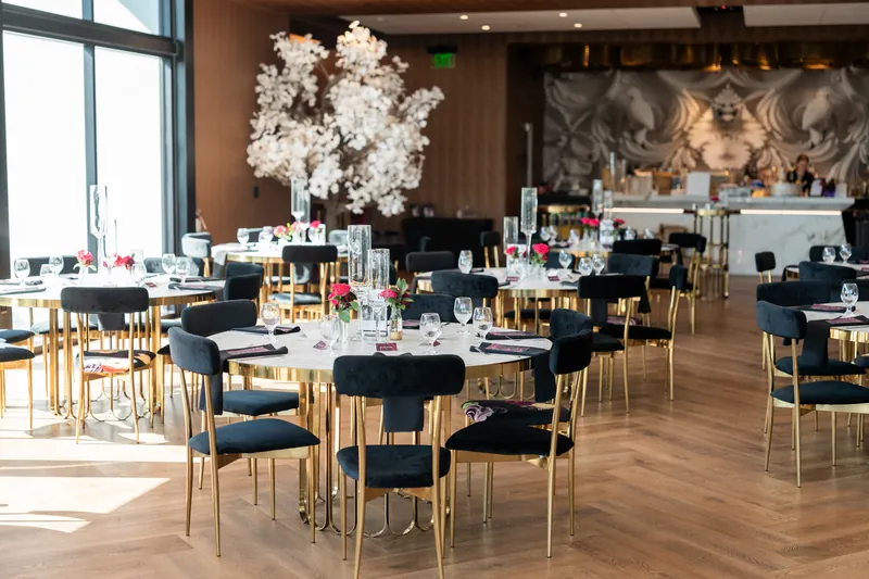Modern reception space with round tables in black linens, gold chairs, and tall white floral centerpieces by large windows