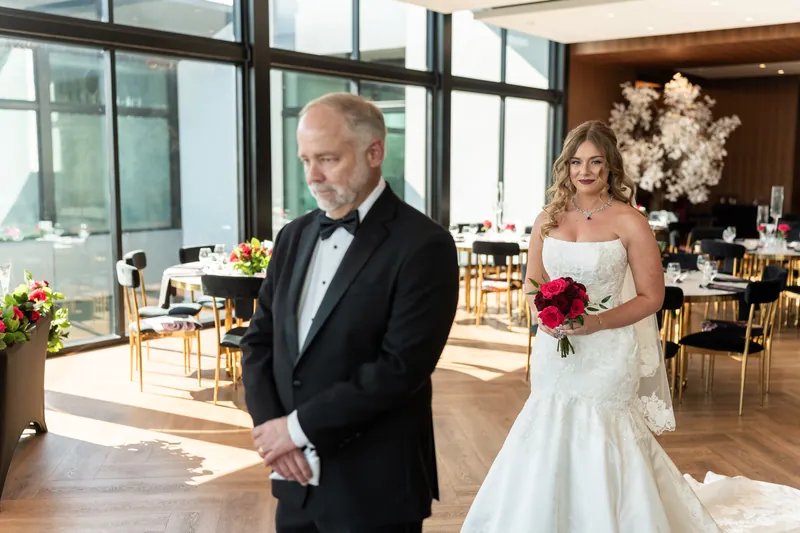 Lauren and Kevin side by side in the reception hall, Kevin in a tuxedo and Lauren in her gown with a red bouquet