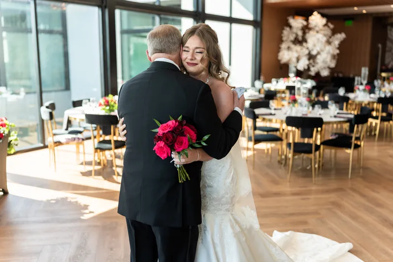 Kevin and Lauren embrace holding a fuchsia bouquet in a sunlit ballroom with gold chairs and white florals behind