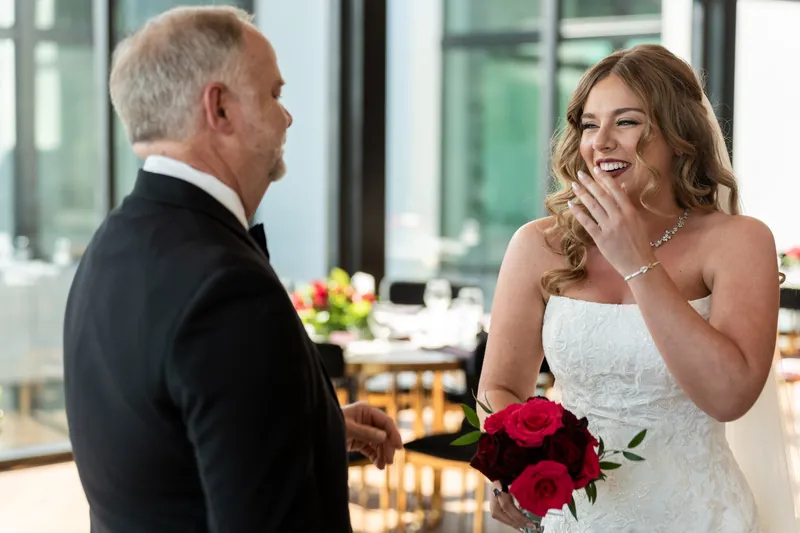 Kevin and Lauren embrace as she laughs while holding her red rose bouquet at their first dance in the reception venue.