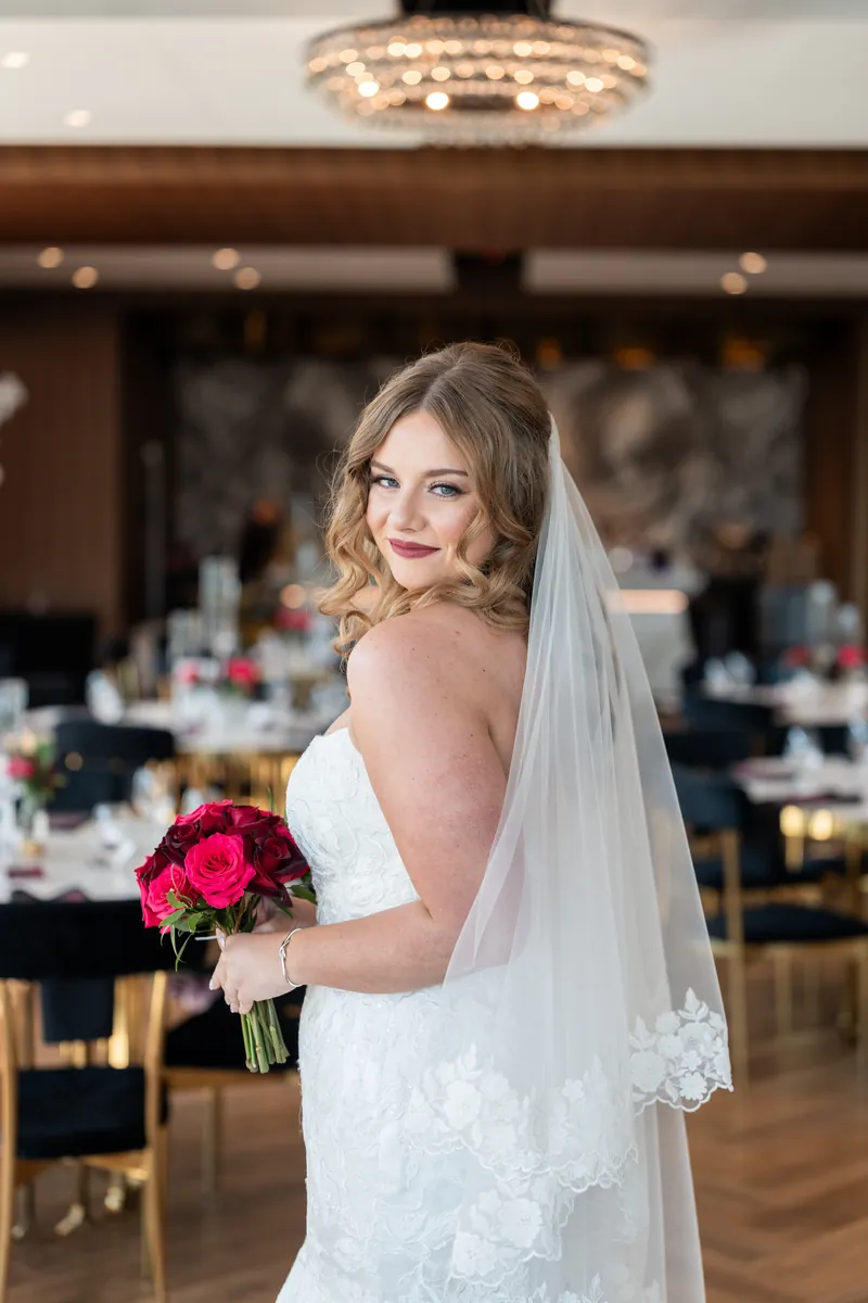 Lauren in her strapless gown and veil, holding her red bouquet and looking over her shoulder in the reception hall