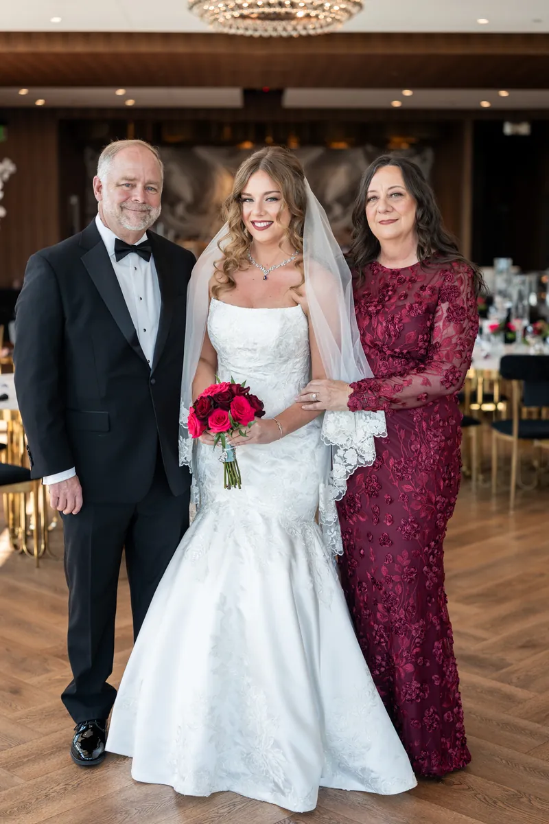Lauren in a white mermaid wedding dress with a red bouquet stands between two guests inside an elegant reception hall.