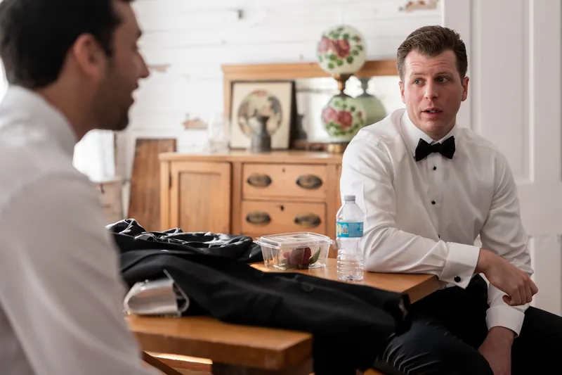 Kevin sits at a wooden dresser wearing a white dress shirt and bow tie while talking with a groomsman in a suit jacket.