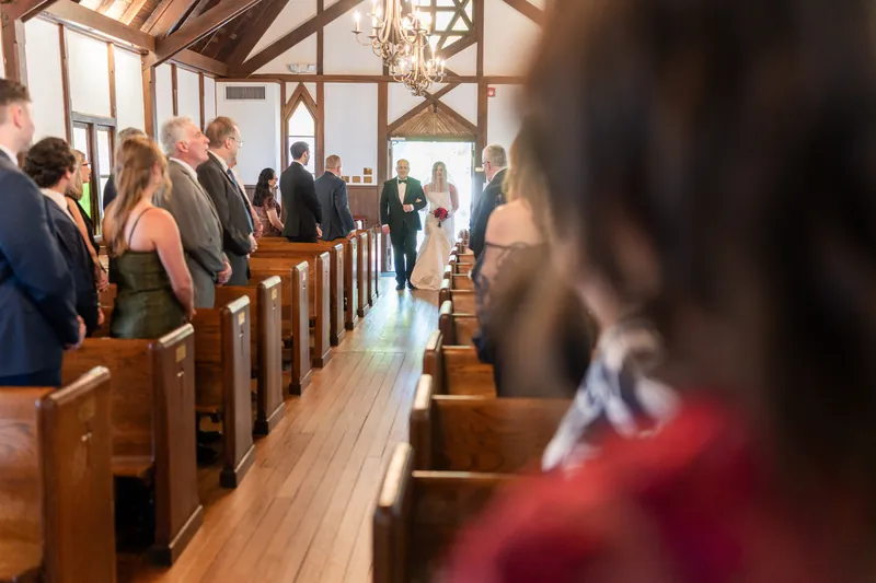 Wedding guests seated in wooden pews fill a bright chapel with large windows overlooking the grounds.