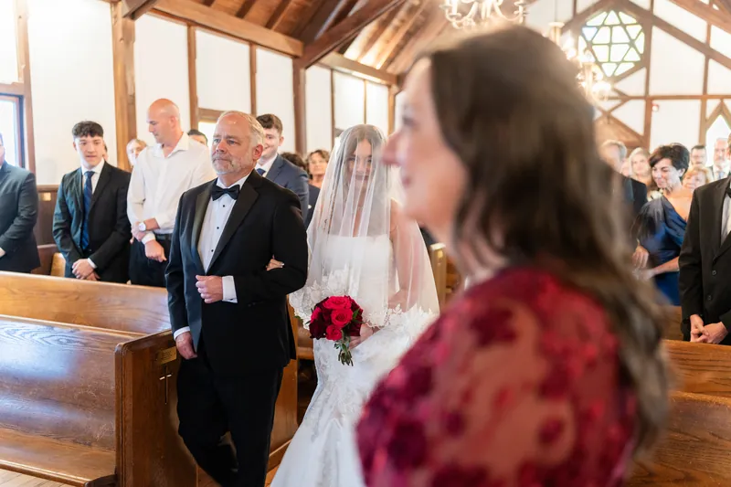 Lauren walks down the aisle in her dress, veil, and red bouquet as Kevin waits in a dark suit in the chapel