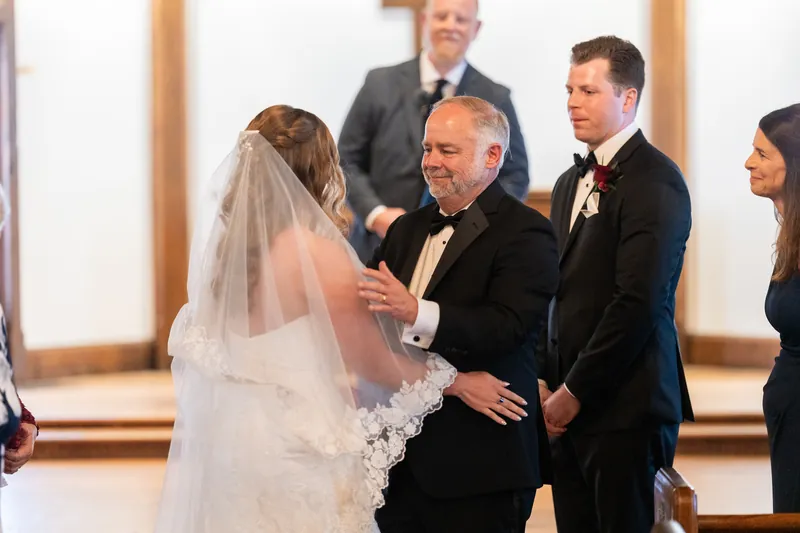Lauren's father places her hand into Kevin's as they stand together in their wedding attire during the ceremony.