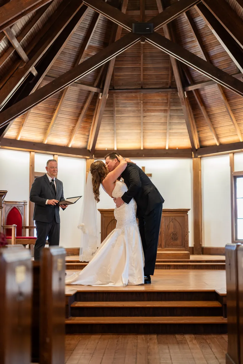 Kevin kisses Lauren after their vows at the altar in the wood-beamed chapel with officiant and guests watching