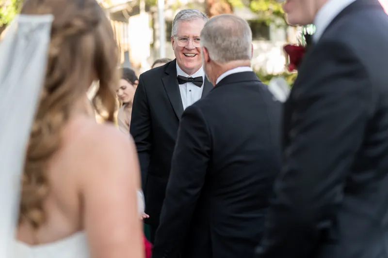 Kevin smiles while greeting guests in bow ties during the reception, with Lauren visible in her white dress.