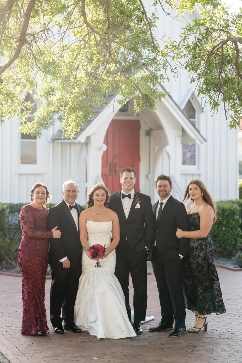 Lauren and Kevin stand with five family members in formal attire in front of the white chapel on a brick pathway.