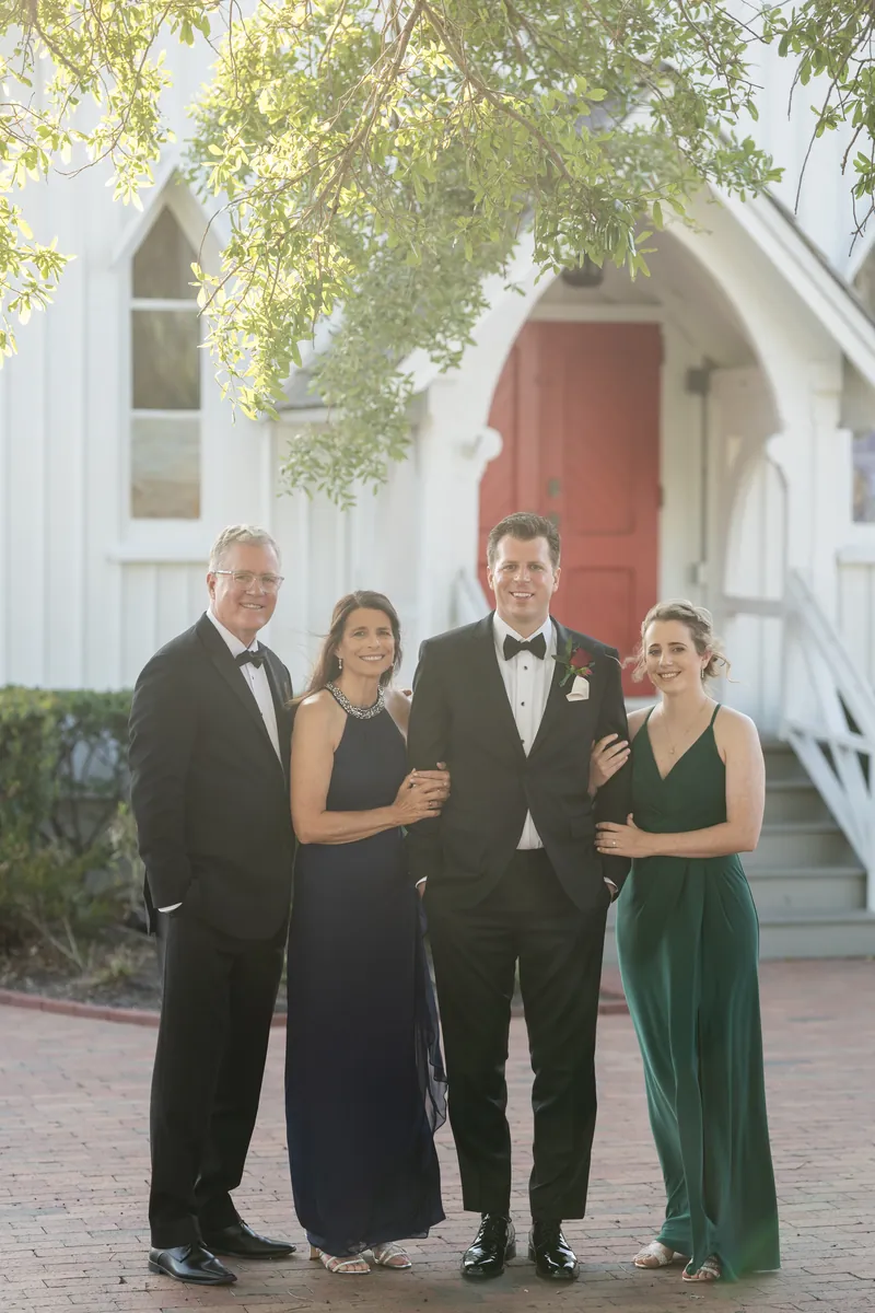 Four formally dressed family members stand together on a brick pathway in front of a white chapel with ivy overhead.