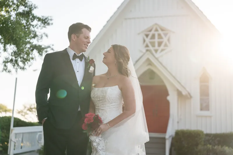 Kevin in a dark suit and Lauren in a strapless white gown stand together with the sunlit chapel visible behind them.