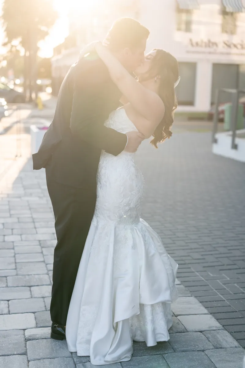 Kevin dips Lauren backward as she holds his shoulders, both smiling on a brick plaza in warm sunlight