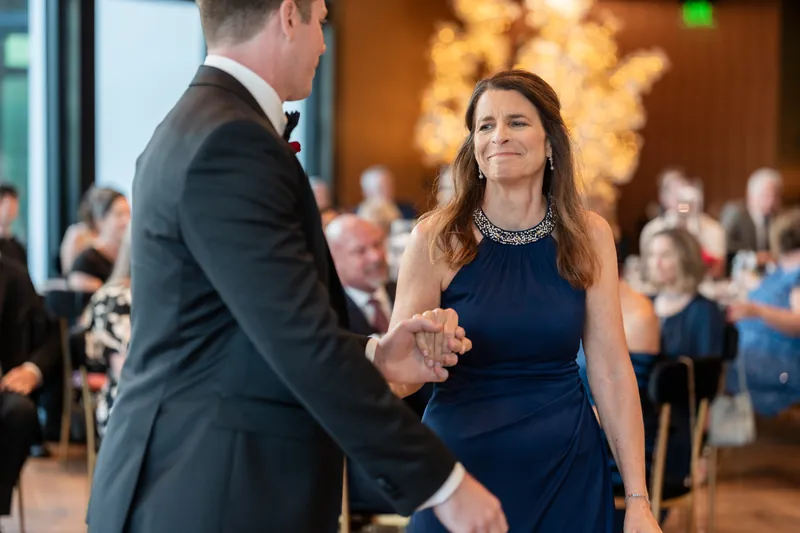 A man and woman hold hands while dancing in a ballroom with warm golden lighting and guests seated in the background.