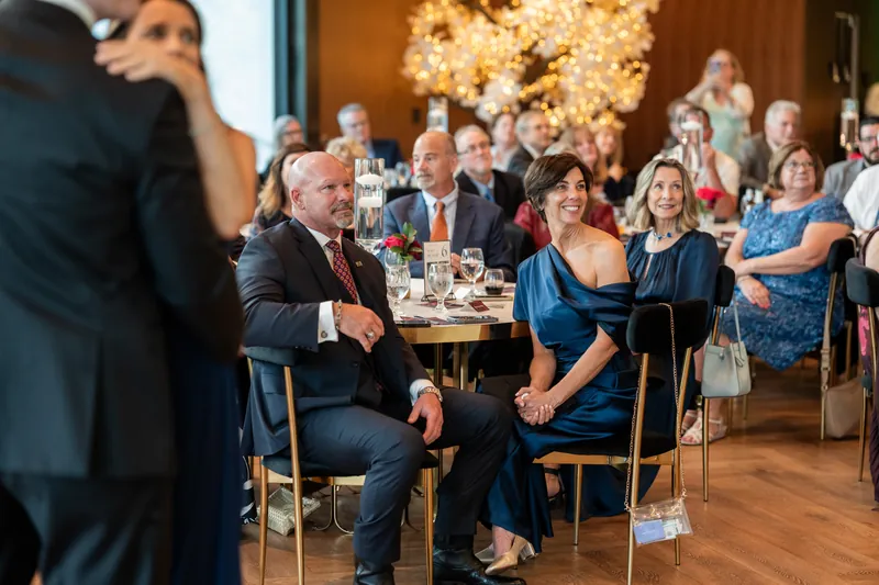 Wedding guests seated at tables watch the reception, with a floral arrangement visible and warm uplighting behind them.