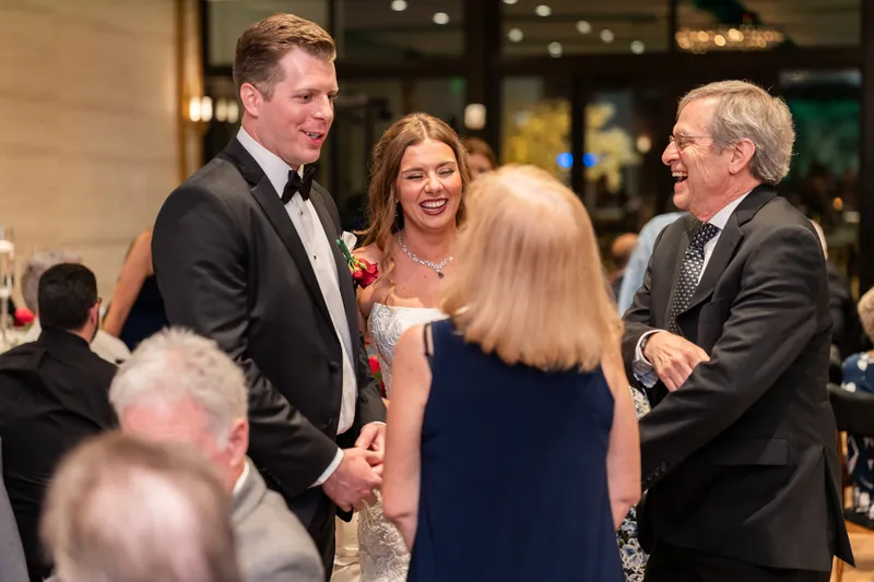 Kevin in a tuxedo chats with guests at the reception, including a woman in a navy dress and an older man in a gray suit.