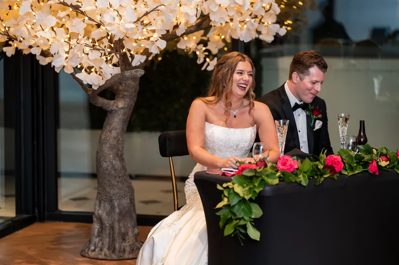 Lauren and Kevin at their table, Lauren in her gown laughing with champagne beneath an illuminated cherry blossom tree