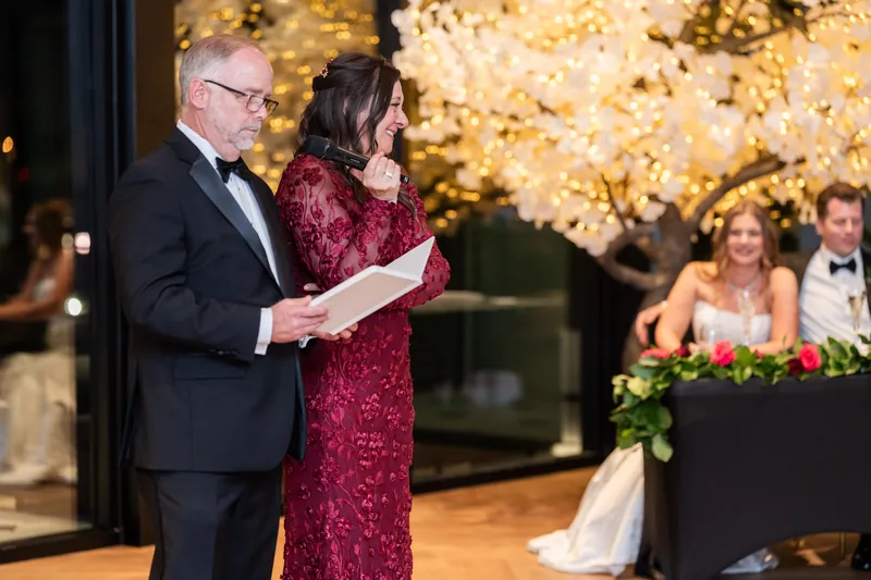 A man in a tuxedo holds papers while standing with a woman in a burgundy dress at the microphone at the evening reception.