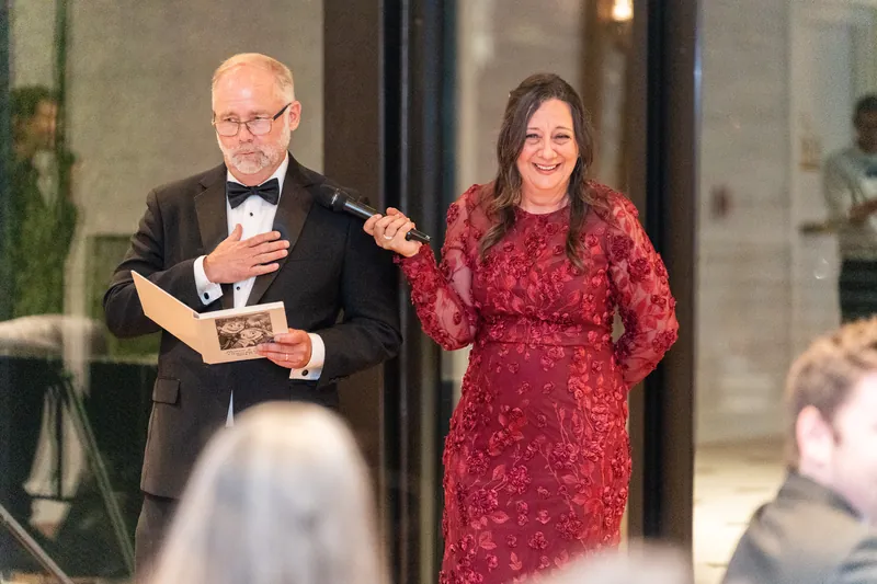 A man in a tuxedo holds a framed photo while standing next to a woman in a red sequined dress during a reception speech.