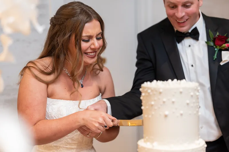 Lauren cuts into a white wedding cake with Kevin in a tuxedo beside her, both smiling.