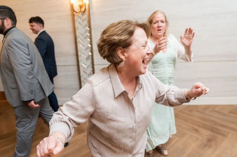 A woman in a beige shirt dances with arms raised while guests watch on a wooden dance floor.