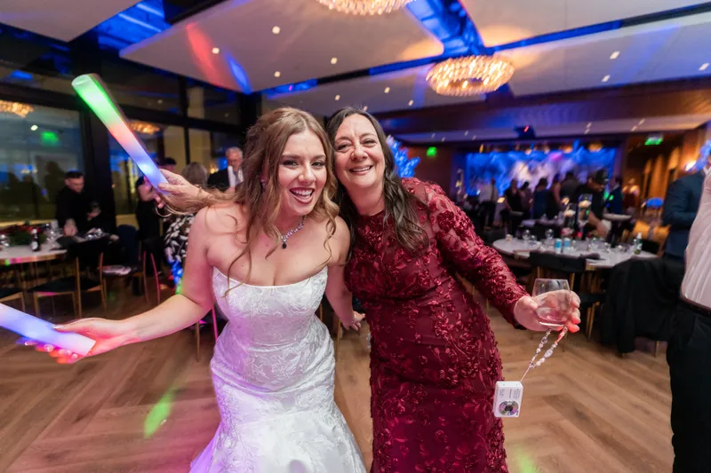 Lauren in her white wedding dress and a woman in a red sequined dress smile together on the dance floor under blue lighting.