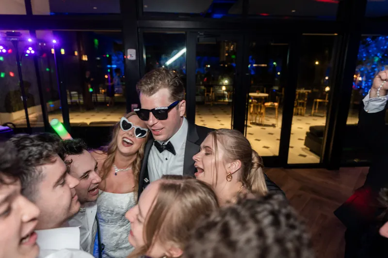 Kevin wearing sunglasses at the reception surrounded by guests, with blue city lights visible through windows behind them.