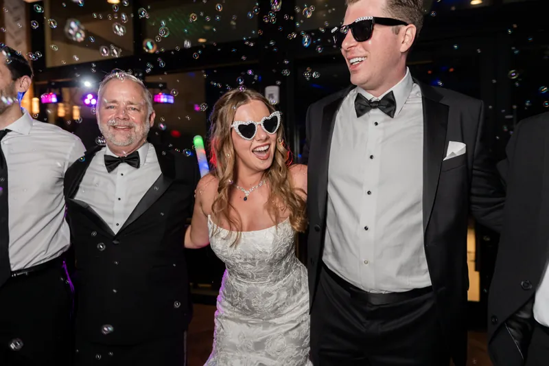 Kevin in a tuxedo and sunglasses standing with a blonde woman in a beaded white dress and other guests during the reception.