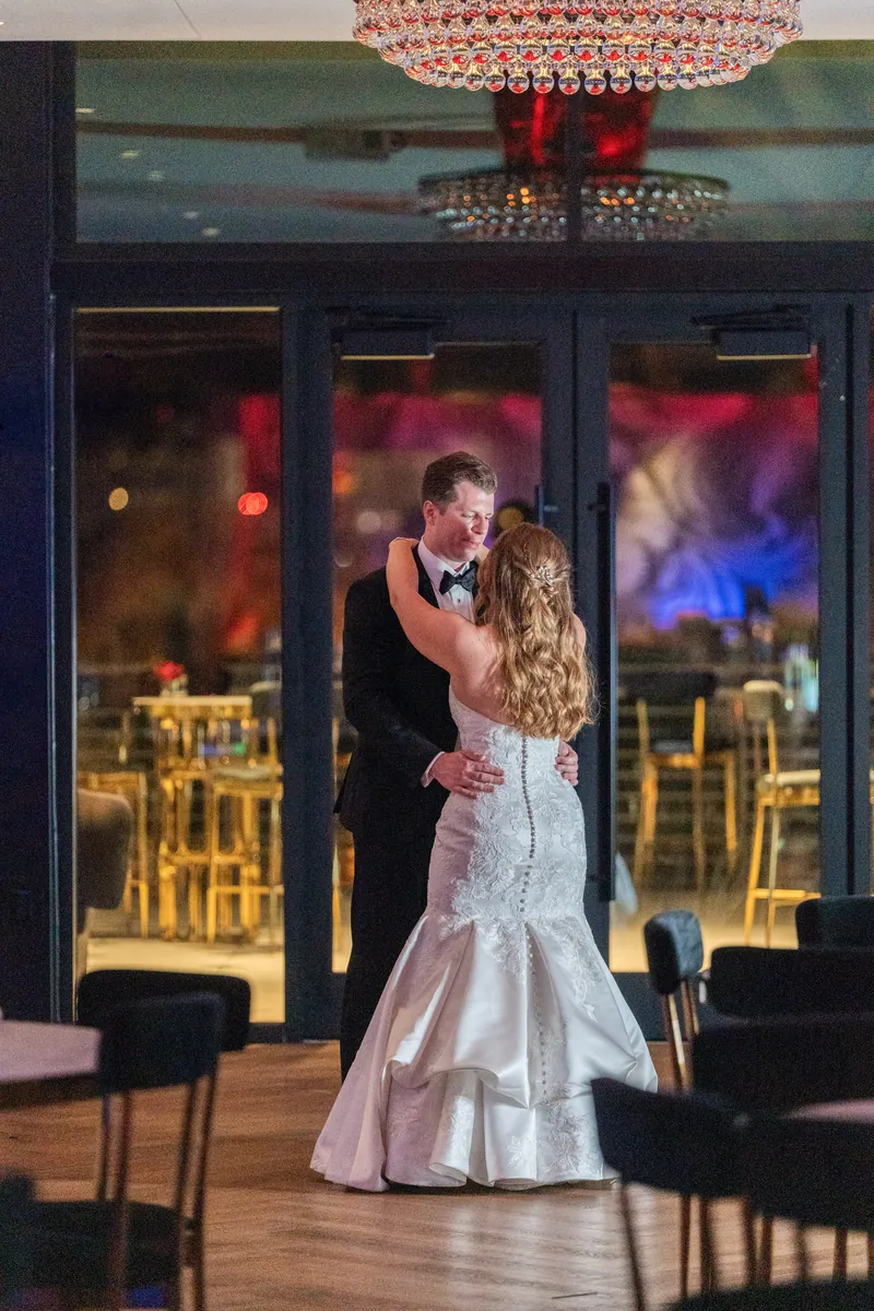 Kevin and Lauren dance under a red beaded chandelier with gold chiavari chairs and blue lighting visible behind them.