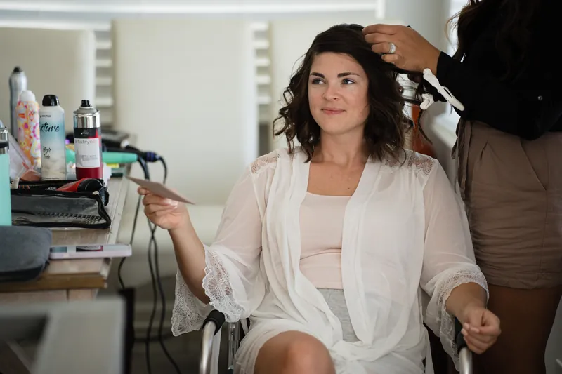 Laurel gets ready in a bridal suite, adjusting her white robe while someone styles her hair at a vanity mirror.