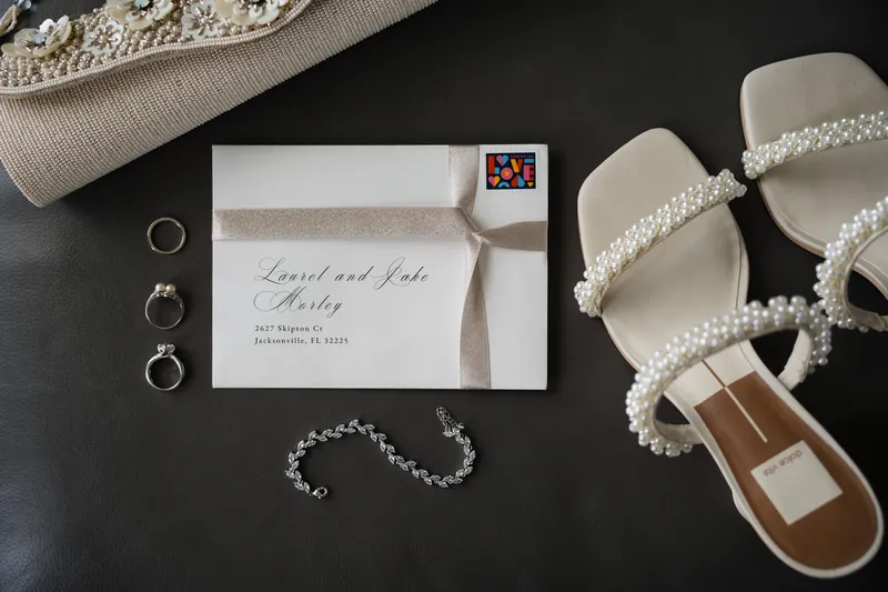 Wedding day details on dark surface: two gold rings, pearl bracelet, beaded shoes, and envelope addressed to Laurel and Jake.