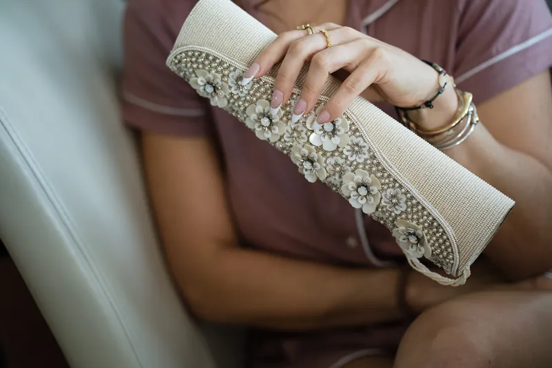 A person in a mauve dress holds a white beaded clutch, displaying pearl bracelets on their wrist near a window.