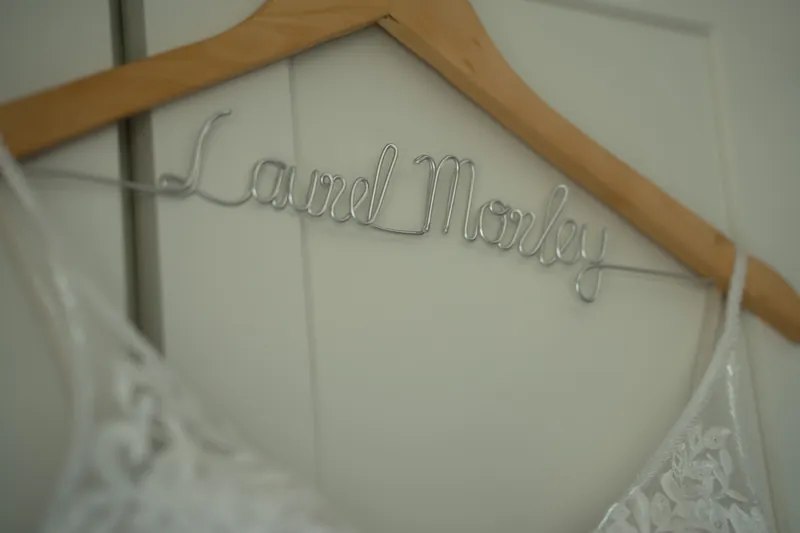 A wooden hanger holds a personalized name tag reading "Laurel & Jake" above a white lace wedding dress.