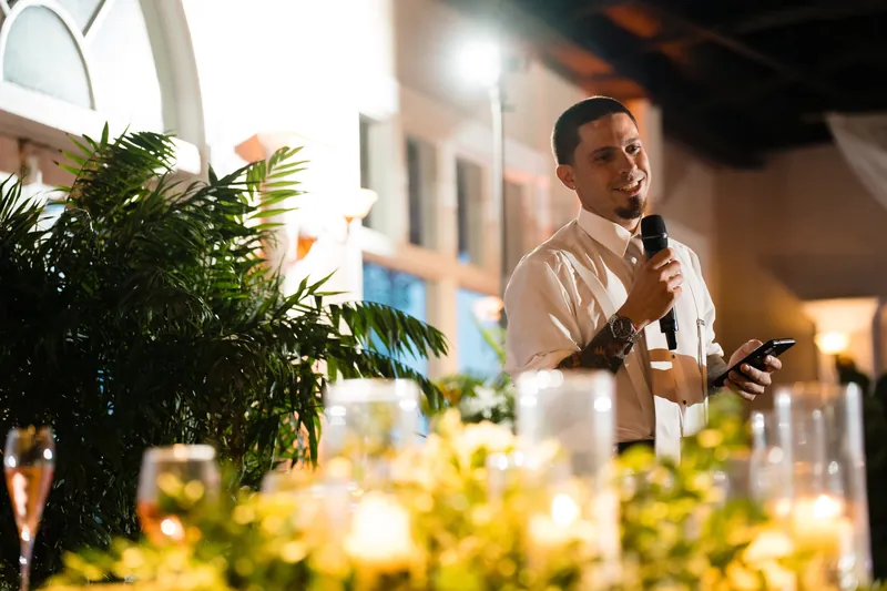Jake holds a microphone during his speech, framed by greenery and ambient golden lighting in an elegant indoor setting.