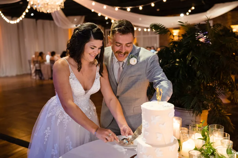 Laurel and Jake smile as they cut their wedding cake together, surrounded by greenery and string lights.