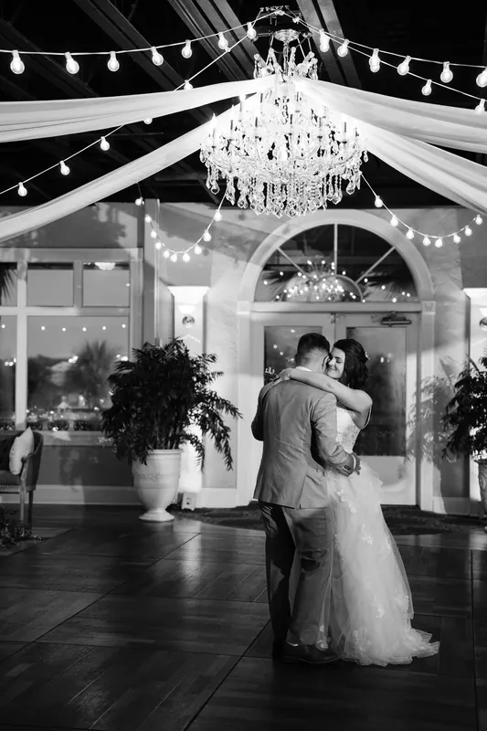 Laurel and Jake dance under a crystal chandelier draped with fabric and twinkling lights in this black-and-white photograph.