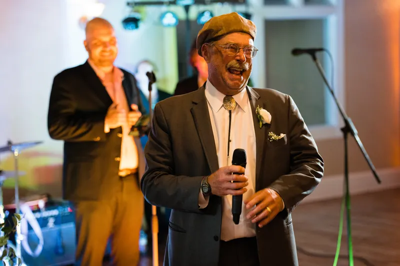 A man in glasses and dark suit speaks at a microphone during the reception at The White Room.