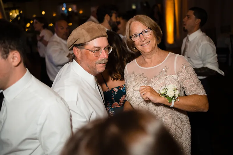An older couple dances together, the man in a cap and glasses, the woman in a beaded champagne dress with a corsage.