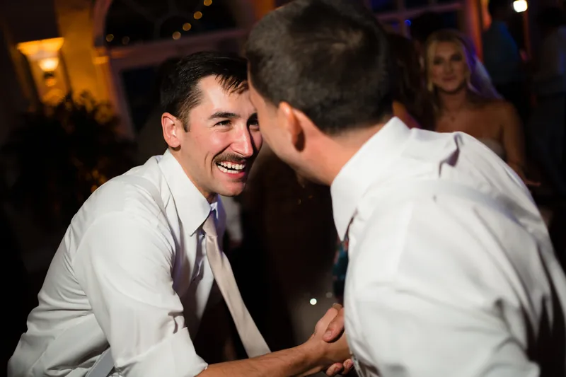 Jake grins widely while dancing face-to-face with another guest in the dimly lit reception space.