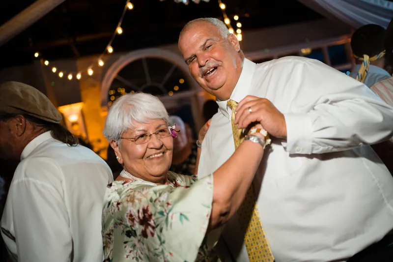 An older man in a white shirt and yellow tie dances with an older woman in a floral dress, both smiling broadly.