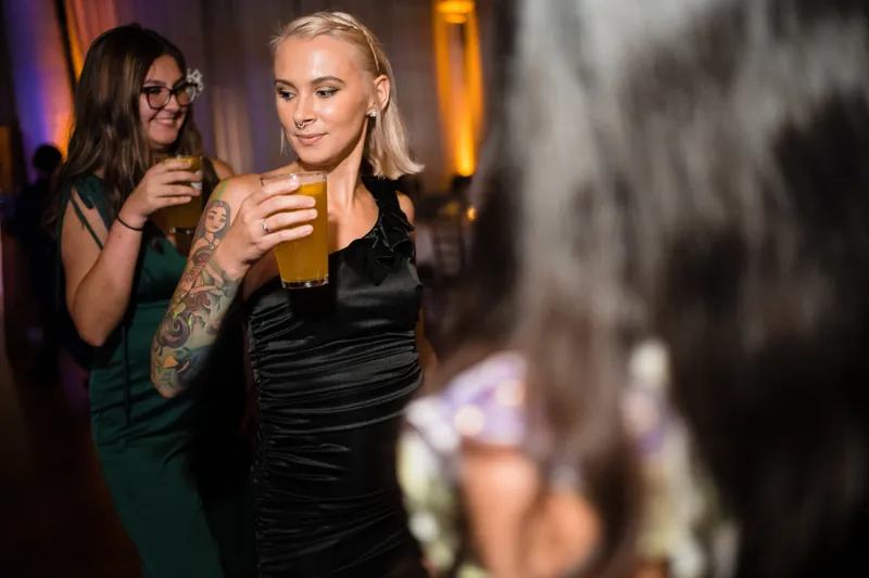 Two guests holding beers laugh together at The White Room, one in a black velvet dress and the other in green.