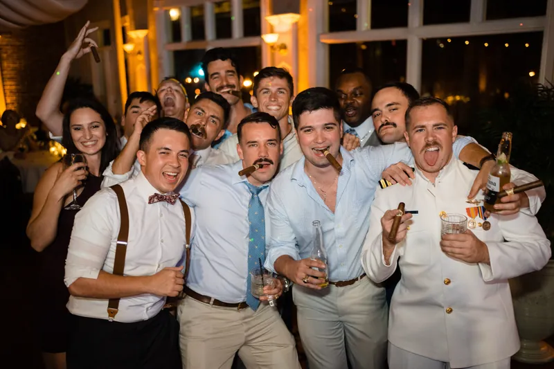 Jake poses with groomsmen in light blue shirts and suspenders, raising drinks in celebration.