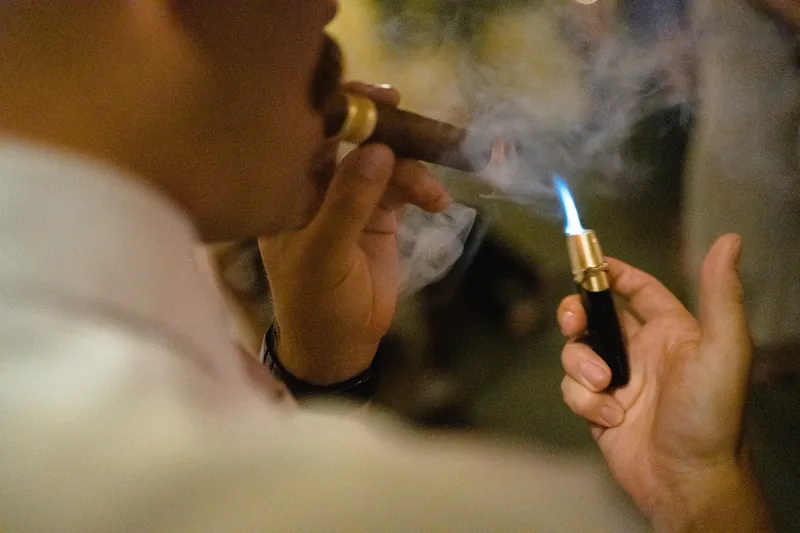 One hand lights a cigar with a blue flame lighter as smoke curls between them.
