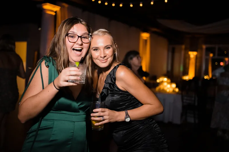 Two female guests in green and black dresses smile and clink glasses indoors.